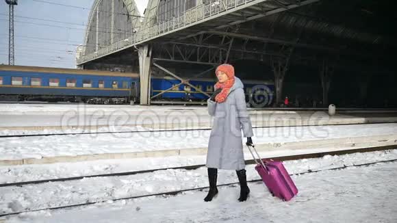 阳光明媚的冬日里一位身穿蓝色外套提着紫色行李箱的年轻快乐女子在火车站视频的预览图