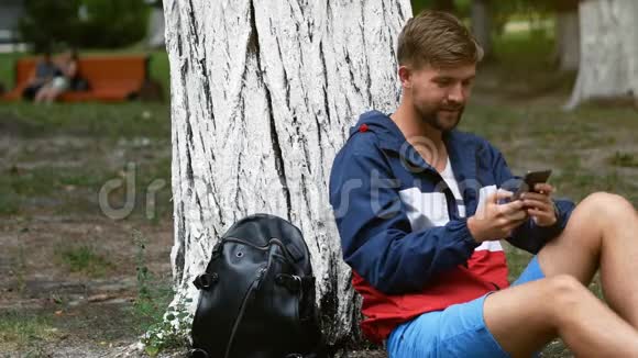 快乐的年轻人在公园里坐在树下用电话交谈户外使用智能手机的大胡子学生视频的预览图