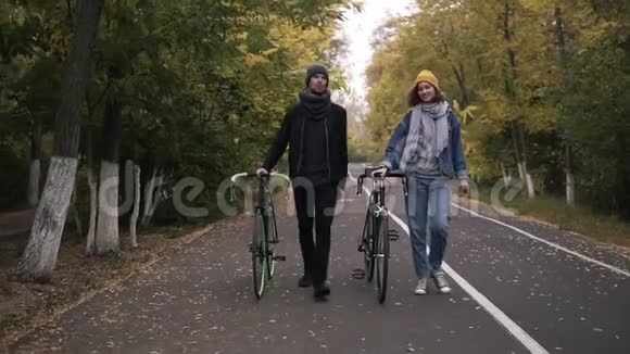 两位亲密的朋友在绿黄高树之间的城市公园里骑着自行车散步聊得很开心视频的预览图