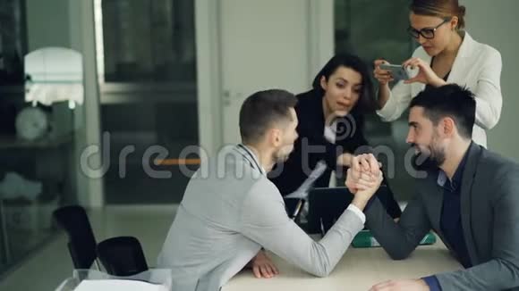 穿西装的帅哥在办公室坐在办公桌前进行手臂摔跤比赛而女孩们则在用视频的预览图