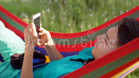 年轻人在花园里的吊床上玩智能手机游戏视频的预览图