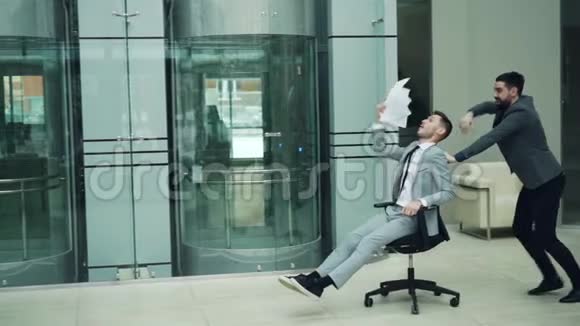 穿着西装的快乐男人慢动作在办公室大厅里玩骑着滚动的椅子扔着文件大喊大叫视频的预览图