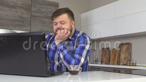 满脸胡子的男人穿着格子衬衫在厨房的笔记本电脑里看新闻喝咖啡动作很慢视频的预览图