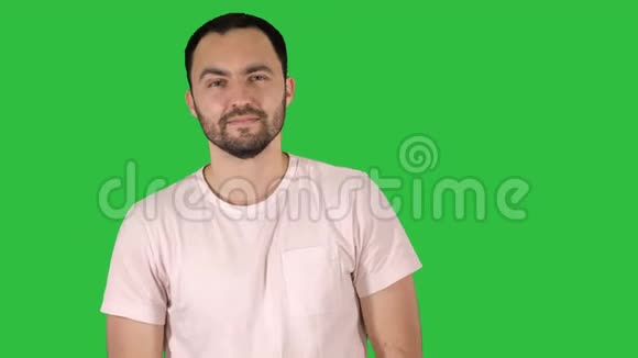 年轻的快乐男人穿着t恤对着镜头微笑走在绿色的屏幕上Chroma键视频的预览图