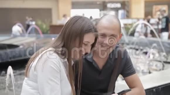 在喷泉背景下坐在购物中心的男人和女孩在电话里讨论照片笑了起来视频的预览图