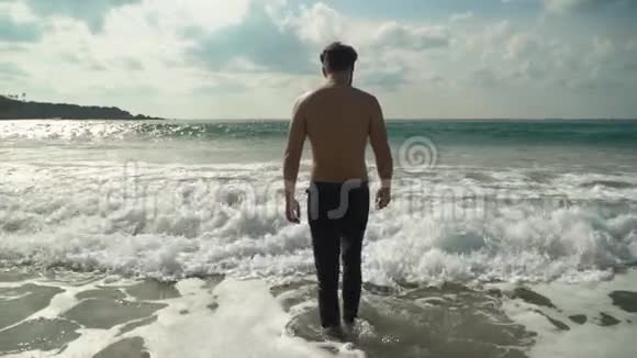 穿牛仔裤的年轻人慢慢地背对着镜头进入浅水海浪在岸上翻滚夏季视频的预览图