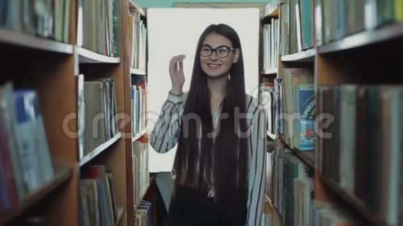 漂亮的年轻女孩留着长长的黑发走到图书馆的书架之间微笑着视频的预览图