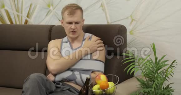 一个手臂骨折的痛苦男人戴着臂架坐在沙发上喝啤酒视频的预览图