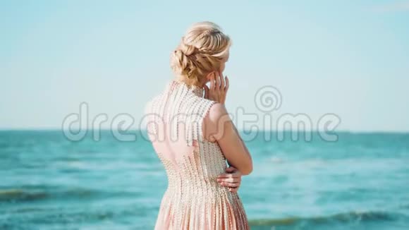 海洋海岸可爱的女人独自站着用手指抚摸她的耳朵用一只手拥抱自己金发视频的预览图