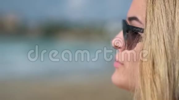 一个戴着深色太阳镜的金发女孩的特写镜头舔着躺在沙滩上的躺椅上美丽的嘴唇视频的预览图