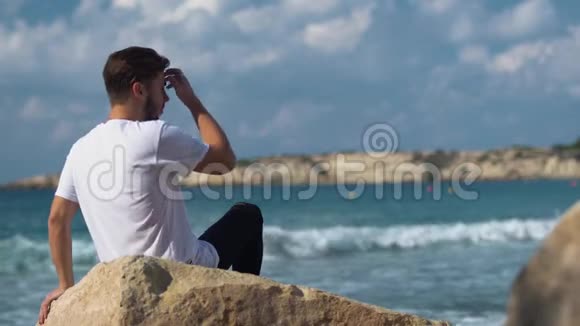 一位年轻的帅哥坐在靠近神奇大海的石头上视频的预览图