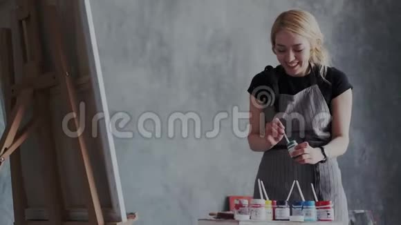 穿着围裙的快乐金发女艺术家在做她喜欢做的事她站在画架前混合颜料这就是视频的预览图