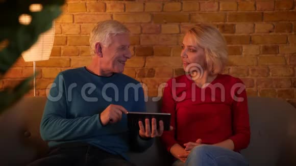 高年级幸福夫妇的特写镜头使用平板电脑愉快地坐在舒适的室内沙发上微笑视频的预览图