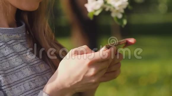 在春城散步的苹果树盛开的枝条背景下漂亮的女人用智能手机短信视频的预览图