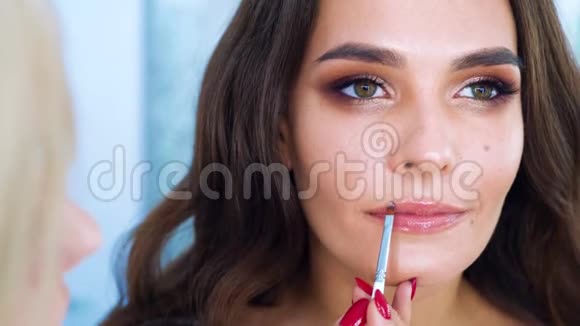特写女化妆师用刷子在年轻的黑发女人的嘴唇上涂口红视频的预览图