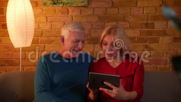 一对年长夫妇在平板电脑上浏览网页的特写镜头幸福地坐在舒适的室内沙发上视频的预览图