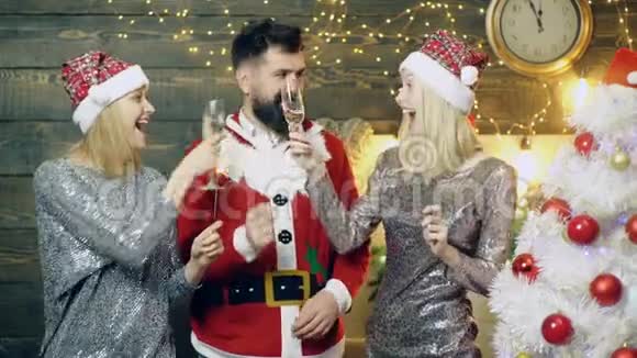戴着圣诞老人服装的大胡子男人和戴着新年帽的两个女孩喝着香槟对着闪闪发光的灯光视频的预览图