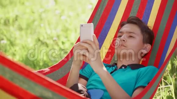 年轻人在花园里的吊床上玩智能手机游戏视频的预览图
