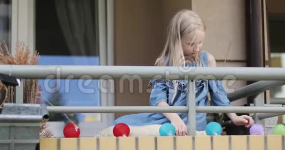 一个穿着连衣裙的可爱金发女孩坐在阳台上玩假日灯视频的预览图