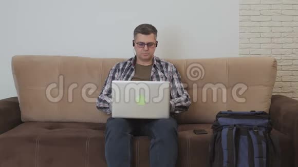 一个中年男子坐在沙发上耳朵里戴着耳机正在使用笔记本电脑旁边是一个大型游客视频的预览图