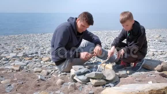 爸爸和儿子在海边手工制作的石头上点燃篝火中的煤家庭周末视频的预览图