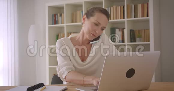 用手提电脑和办公室打电话的老白种人黑发女商人的特写照片视频的预览图