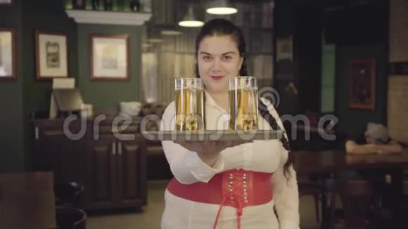梳着辫子的女人穿着白色衬衫拿着托盘拿着两个啤酒杯对着镜头微笑在酒吧休闲视频的预览图