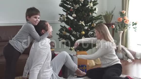 妈妈和孩子把圣诞礼物送给坐在圣诞树旁地板上的父亲幸福的年轻家庭视频的预览图