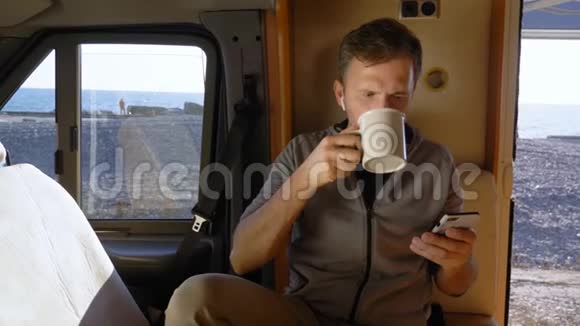 男人喝咖啡坐在海边的大篷车里用智能手机视频的预览图