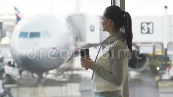 穿运动服的漂亮女孩喝机场附近的外卖咖啡背景是飞机视频的预览图