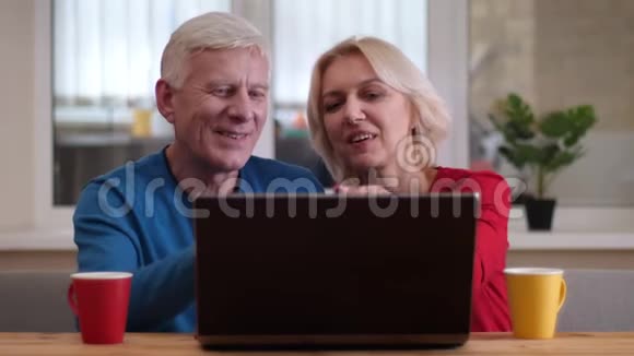 在舒适的室内书桌上放着茶杯的笔记本电脑上播放视频通话的老年幸福夫妇的特写镜头视频的预览图