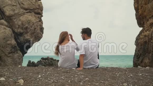年轻的一对夫妇坐在两块石头之间的美丽海滩上交谈着视频的预览图
