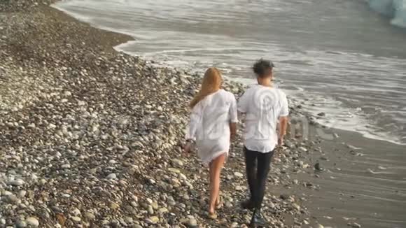 幸福的一对穿着白色连衣裙的夫妇牵着手沿着岩石海岸散步年轻家庭的休闲无人驾驶飞机射击视频的预览图