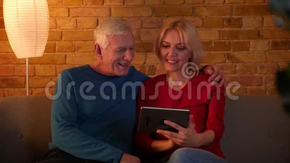 在平板电脑上浏览网页的高级幸福夫妇的特写镜头欢快地坐在室内的沙发上视频的预览图