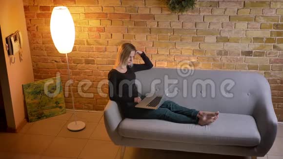 在舒适的室内沙发上坐着时年轻漂亮的女青少年在笔记本电脑上浏览的侧视图视频的预览图