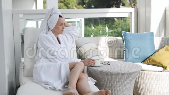美丽的女人穿着浴袍躺在露台上的阳光休息室里喝着咖啡在新鲜空气中放松视频的预览图