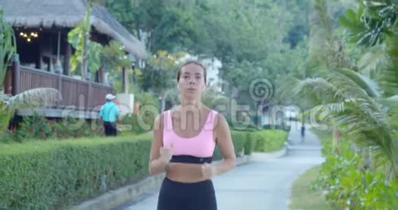 穿着粉色运动上衣的年轻美女沿着热带村庄奔跑复制空间视频的预览图