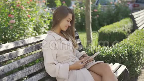 微笑的年轻女子穿着浴袍坐在公园的长凳上检查平板电脑上的信息自信的女孩喜欢视频的预览图