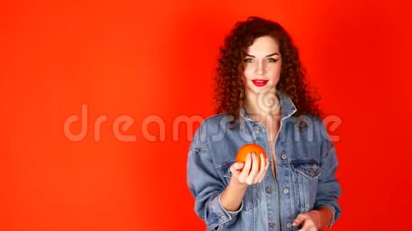 年轻的卷发女孩在工作室的珊瑚背景上扔橙色视频的预览图