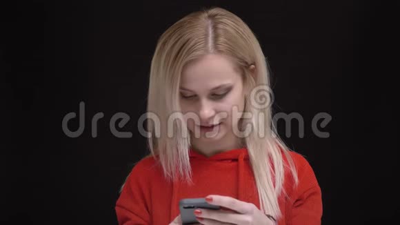 年轻的白发白种人穿着红色毛衣的白种人女孩在黑色背景下微笑地看着智能手机的肖像视频的预览图