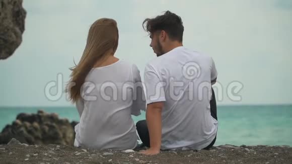 后景年轻漂亮的夫妇坐在海边温暖的鹅卵石上他们愉快地交流一起度过时光视频的预览图
