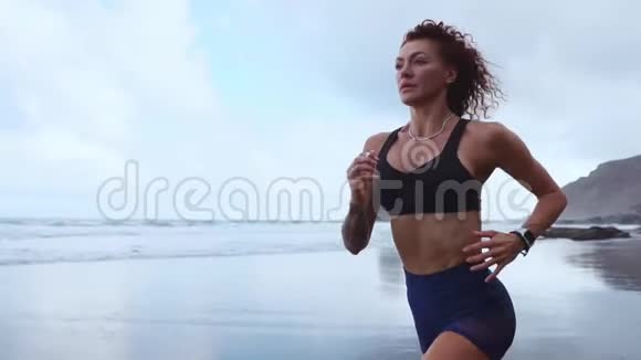 运动女性在海滩上奔跑黑色的沙子在山脉和海洋的背景下缓慢运动关门视频的预览图