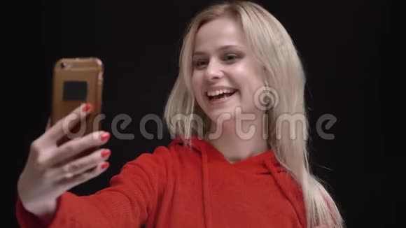 年轻的白发白种人白种人女孩戴着黄色太阳镜微笑着在黑色背景下的智能手机上用视频聊天视频的预览图