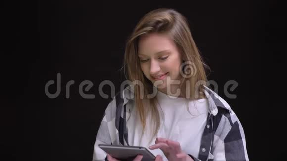 一幅金发白种人长发女孩穿着格子衬衫的肖像在黑色背景上快乐地展示了绿色的平板电脑屏幕视频的预览图