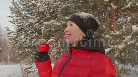 穿着红色冬衣的微笑女人在霜冻的日子在雪林里喝茶视频的预览图