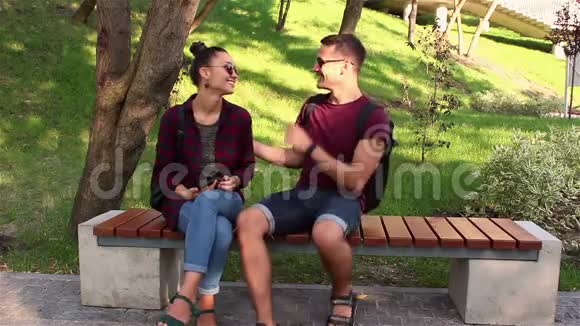 年轻的恋人们在一个阳光明媚的日子里坐在公园的长凳上正在积极地讨论一款新的智能手机视频的预览图