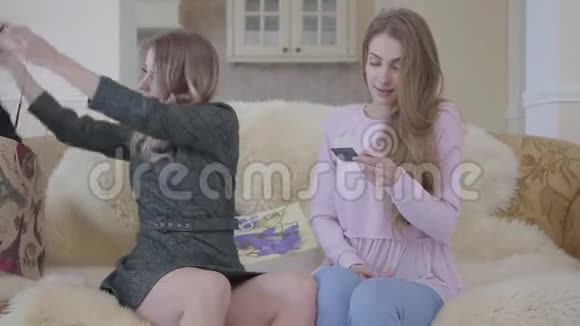 两个时髦的女孩在看购物从购物袋里得到女士们坐在舒适的沙发上青年视频的预览图