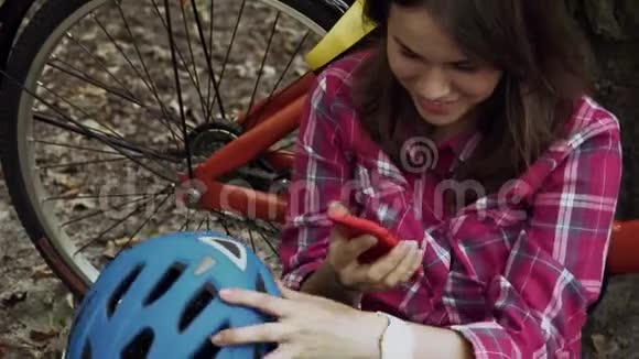 生态自行车运输年轻的高加索女学生坐在公园里休息附近租着橙色自行车用手机视频的预览图