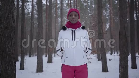 美国黑人女孩穿着温暖的衣服戴着豆帽和夹克站在冬天的森林里看着镜头美丽美丽视频的预览图