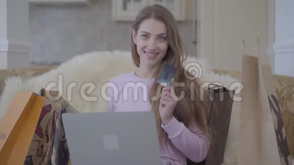 可爱的女人坐在沙发上旁边有一台笔记本电脑和很多购物袋显示着一张黑色信用卡女孩买的视频的预览图